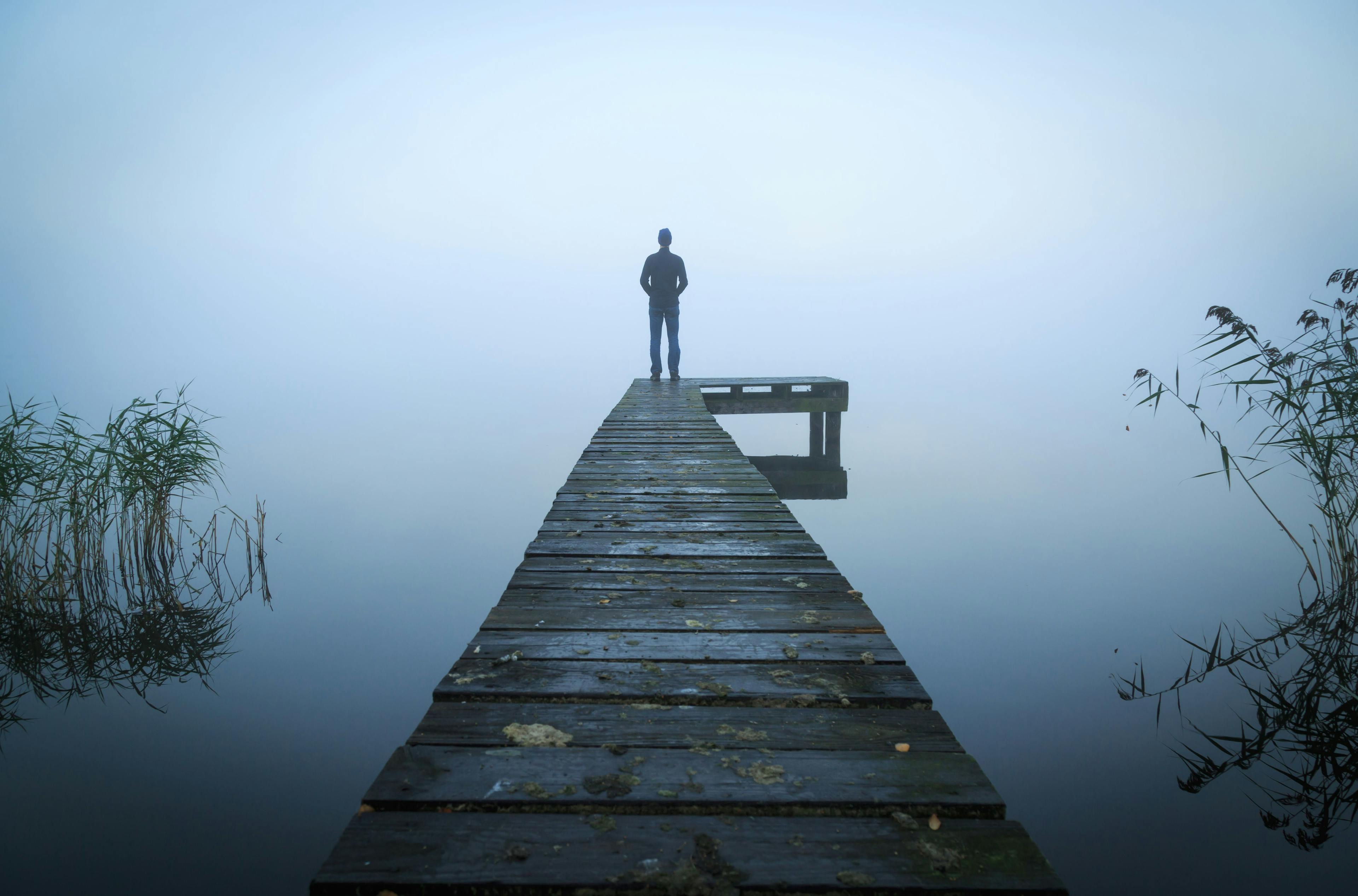 Man standing on a jetty at a lake during a foggy, gray morning/sanderstock/Adobestock